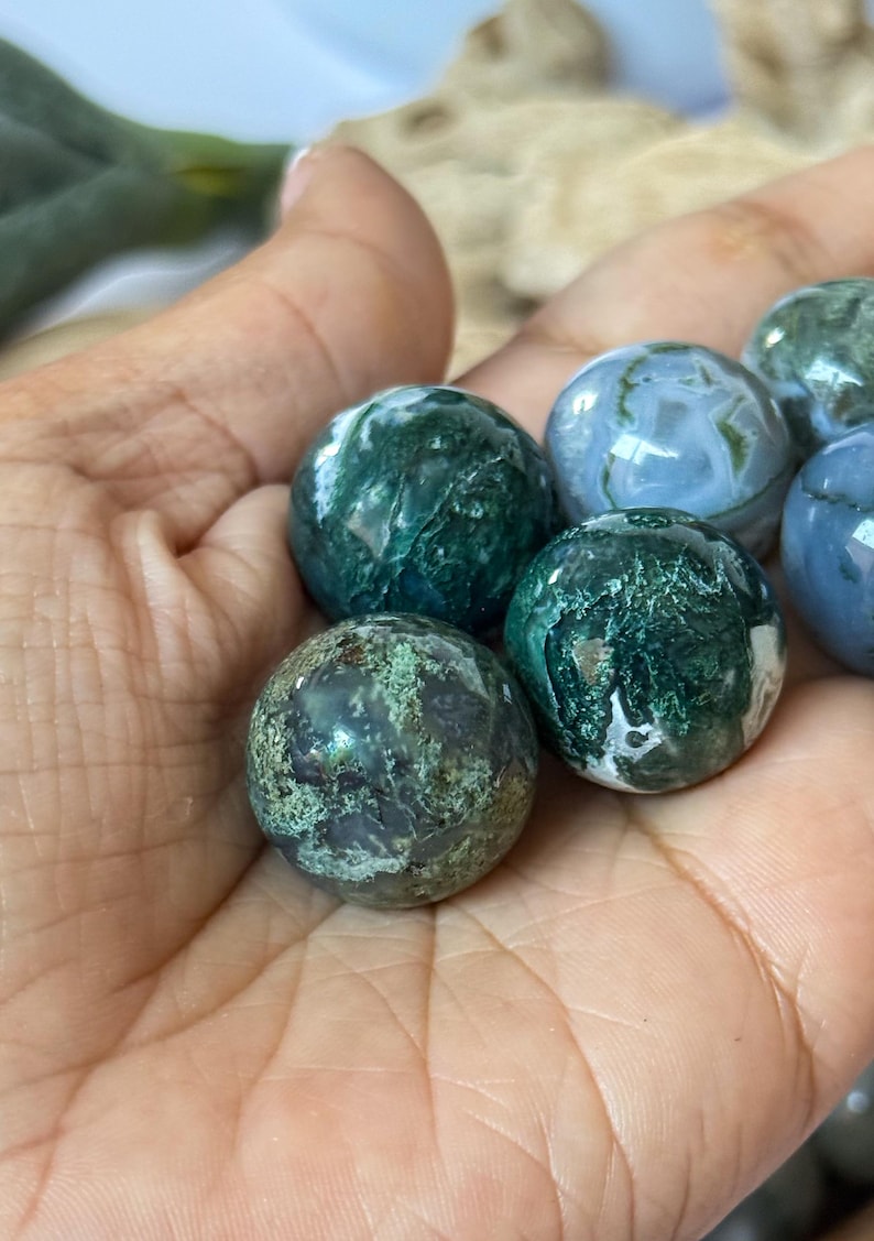 May include: A collection of polished moss agate spheres in varying shades of green and white. The spheres are held in a hand, showcasing their natural patterns and textures. The gemstones have a smooth, rounded shape.