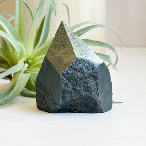 May include: A dark green, faceted, crystal point with a rough, natural texture. The crystal is sitting on a white surface.