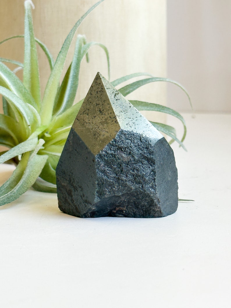 May include: A dark green, faceted, crystal point with a rough, natural texture. The crystal is sitting on a white surface.