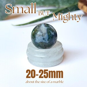 May include: A small, round, moss agate gemstone, approximately 0.79 to 0.98 inches in diameter, rests on a clear, circular stand. The image includes the text "Small BUT Mighty" and "20-25mm about the size of a marble."