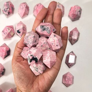 May include: A hand holds a collection of faceted, pink rhodonite crystals. The crystals have a mix of pink and black coloration, with a geometric shape. Several other crystals are scattered on a white surface.