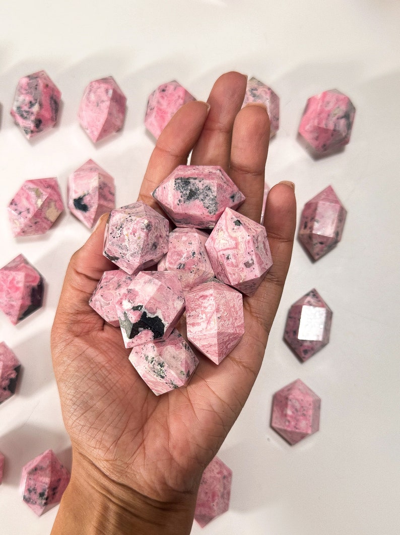 May include: A hand holds a collection of faceted, pink rhodonite crystals. The crystals have a mix of pink and black coloration, with a geometric shape. Several other crystals are scattered on a white surface.