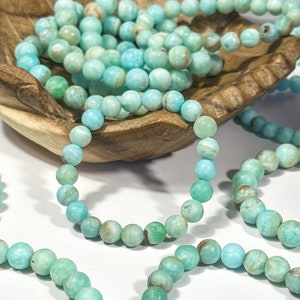 May include: Several strands of turquoise-colored beaded bracelets. The beads are round and have variations in color, ranging from light blue to green. Some bracelets are arranged in a wooden bowl, while others are spread out.