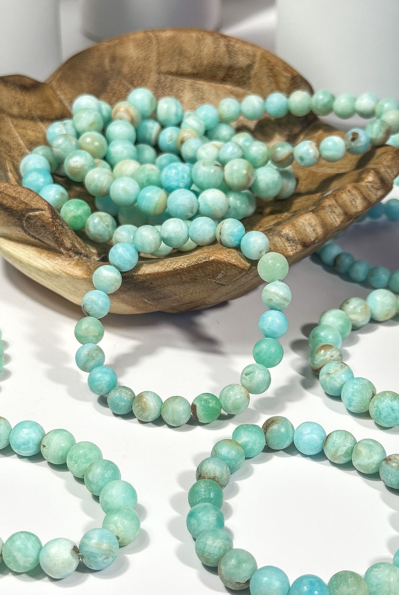 May include: Several strands of turquoise-colored beaded bracelets. The beads are round and have variations in color, ranging from light blue to green. Some bracelets are arranged in a wooden bowl, while others are spread out.