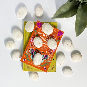 May include: A book titled "The Witch's Guide to Esta" with a colorful cover featuring butterflies, flowers, and stars. The book is surrounded by white stones on a white surface.