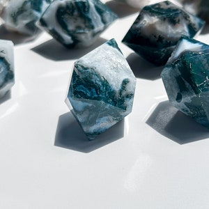 May include: Several faceted, diamond-shaped crystals with a marbled effect. The crystals display a mix of white and deep teal-green hues, giving a natural, organic look. The crystals are arranged on a white surface.