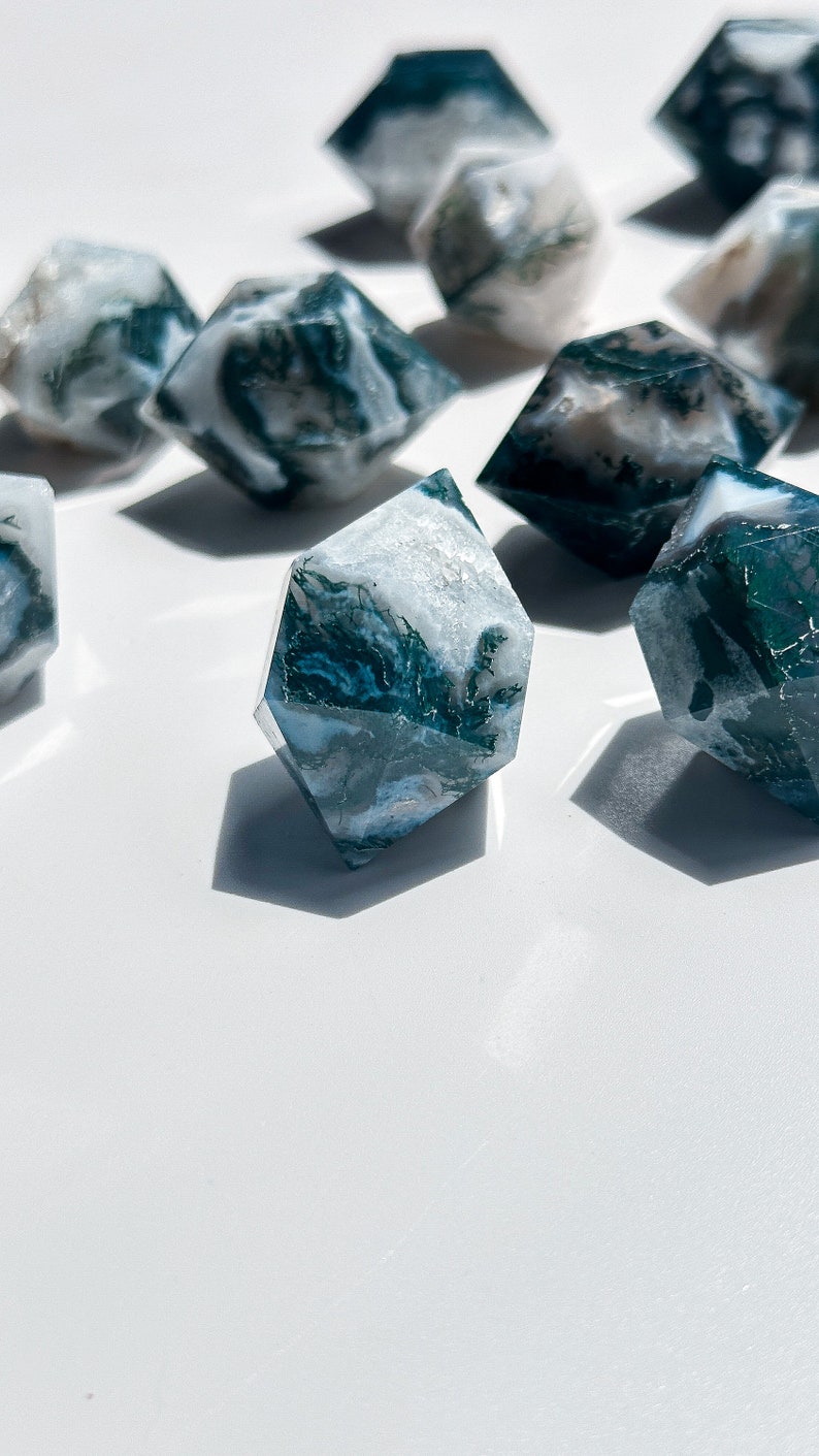 May include: Several faceted, diamond-shaped crystals with a marbled effect. The crystals display a mix of white and deep teal-green hues, giving a natural, organic look. The crystals are arranged on a white surface.