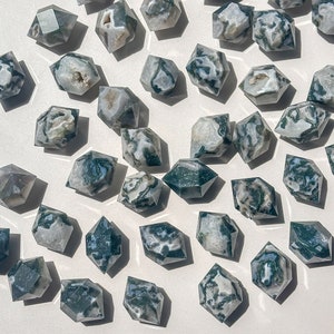 May include: A selection of faceted moss agate crystals. Each crystal displays a unique pattern of green, white, and translucent areas. The crystals are arranged on a white surface, with natural light creating shadows.