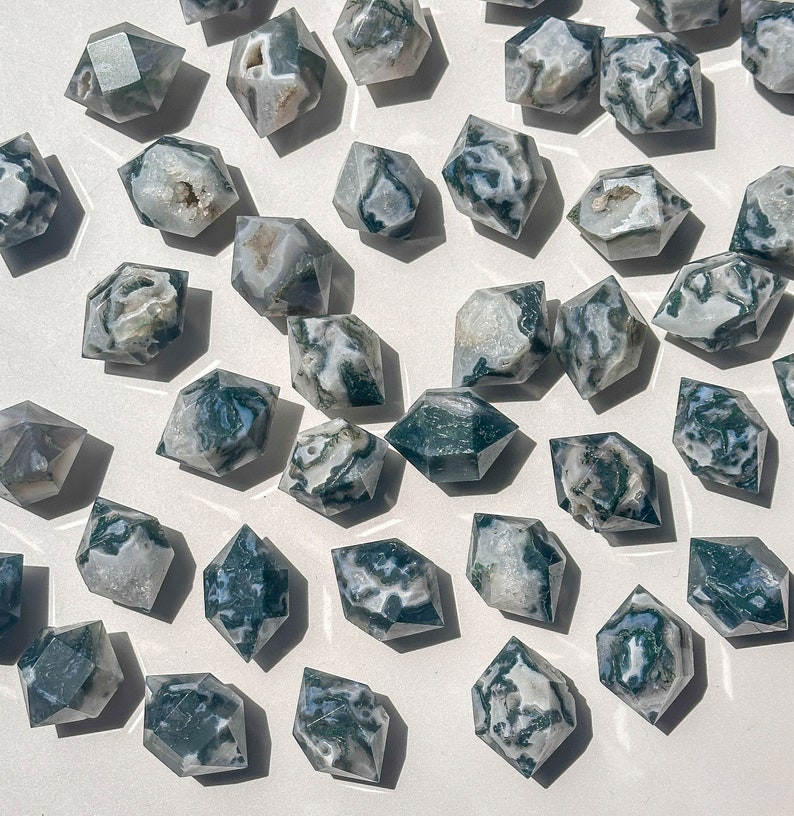 May include: A selection of faceted moss agate crystals. Each crystal displays a unique pattern of green, white, and translucent areas. The crystals are arranged on a white surface, with natural light creating shadows.