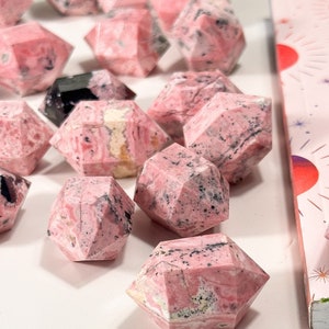 May include: Pink and black faceted gemstone crystals. The crystals have a geometric shape and are arranged on a white surface. The crystals have a pink base colour with black and white veining.