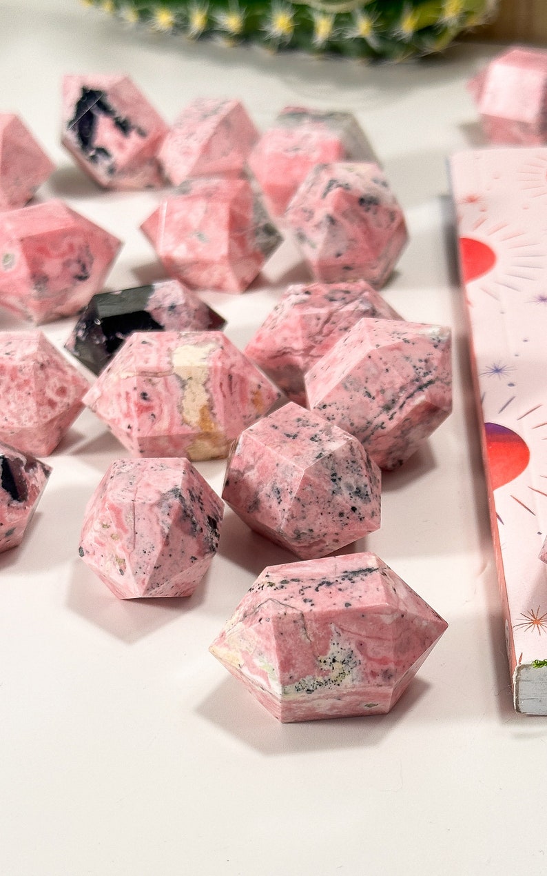 May include: Pink and black faceted gemstone crystals. The crystals have a geometric shape and are arranged on a white surface. The crystals have a pink base colour with black and white veining.