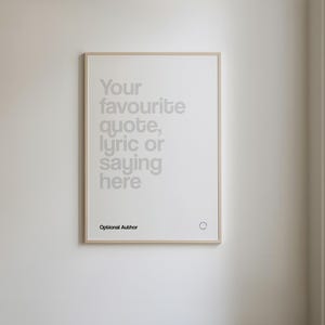 May include: A framed print with a light wood-coloured frame hangs on a white wall. The print features the text "Your favourite quote, lyric or saying here" in a light grey font. Below the text is "Optional Author".