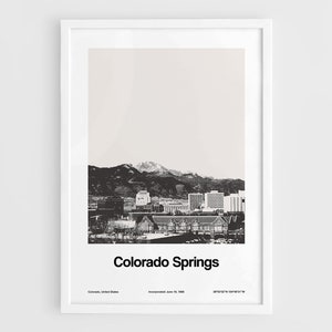 Puede incluir: Una fotografía en blanco y negro de Colorado Springs, Colorado. La imagen muestra el horizonte de la ciudad con Pikes Peak en el fondo. El texto "Colorado Springs" está impreso debajo de la imagen, junto con la fecha de incorporación de la ciudad y sus coordenadas.
