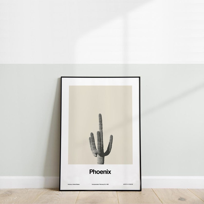 May include: A black framed poster with a beige background featuring a black and white illustration of a saguaro cactus. The text "Phoenix" is printed in black at the bottom of the poster. The text "Arizona, United States" and "Incorporated February 25, 1881" are printed in smaller font below the city name.