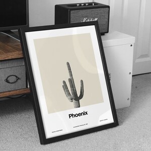 May include: A black framed print with a beige background featuring a black and white illustration of a saguaro cactus. The text "Phoenix" is printed below the cactus. The text "Arizona, United States" and "Incorporated February 25, 1881" is printed below the city name.
