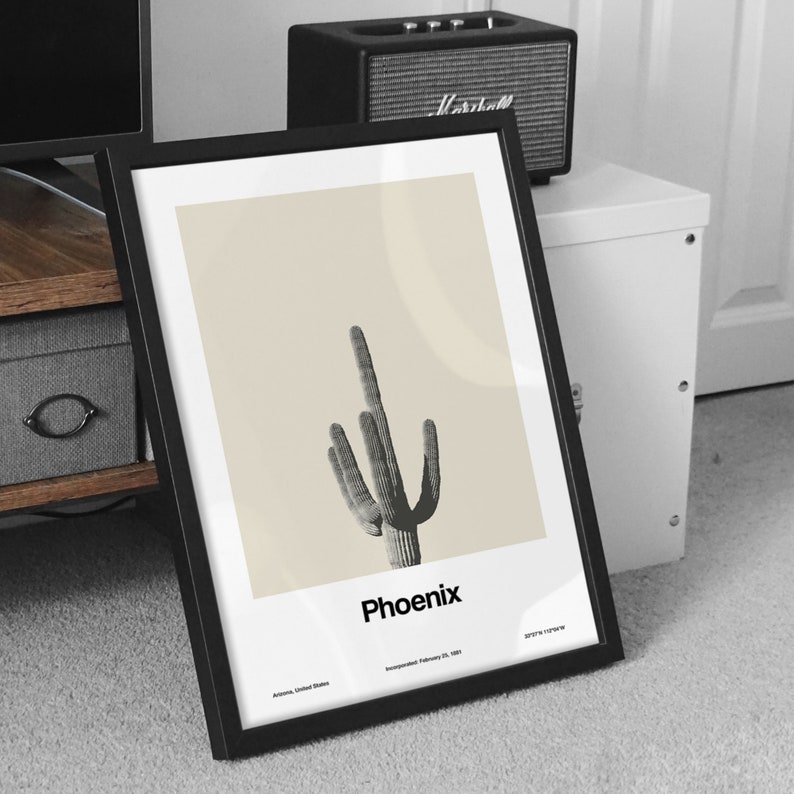 May include: A black framed print with a beige background featuring a black and white illustration of a saguaro cactus. The text "Phoenix" is printed below the cactus. The text "Arizona, United States" and "Incorporated February 25, 1881" is printed below the city name.