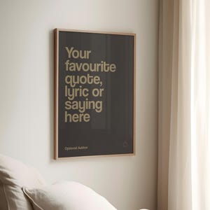 May include: A framed poster with the text "Your favourite quote, lyric or saying here" in a light beige font on a dark grey background. The poster is in a light brown frame and is hanging on a white wall.