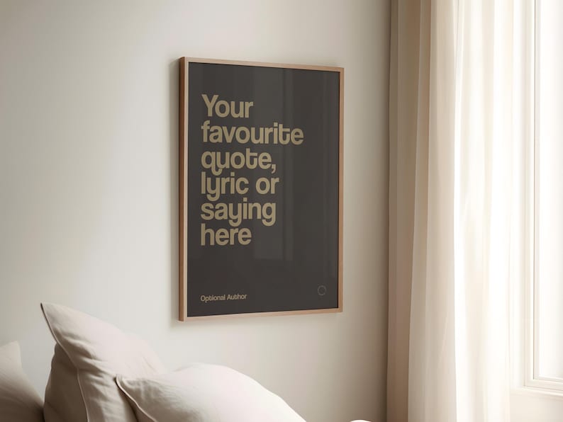 May include: A framed poster with the text "Your favourite quote, lyric or saying here" in a light beige font on a dark grey background. The poster is in a light brown frame and is hanging on a white wall.