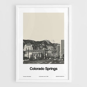 Puede incluir: Una fotografía en blanco y negro de Colorado Springs, Colorado. La imagen muestra una ciudad con edificios y montañas en el fondo. El texto "Colorado Springs" está impreso debajo de la imagen.