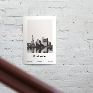 Op de afbeelding: Zwart-witfoto van de skyline van Providence, Rhode Island. De skyline van de stad wordt weerspiegeld in het water. De tekst "Providence" is onder de afbeelding gedrukt.