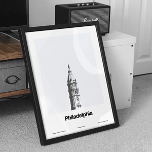 Op de afbeelding: Een zwart-wit ingelijste print van het Philadelphia City Hall gebouw met de tekst "Philadelphia" in vetgedrukte, zwarte letters. De print bevat ook de tekst "Pennsylvania, Verenigde Staten" en "Gesticht 1822".