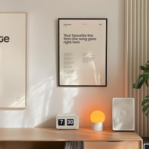 May include: A modern interior scene featuring framed art prints with text, a white digital clock displaying 7:30, a small round lamp, and a white speaker. The artwork includes song lyrics and quotes.