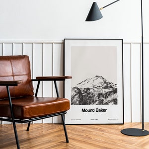 Op de afbeelding: Een zwart-witfoto van Mount Baker, een berg in de staat Washington, is ingelijst en hangt aan een witte muur. Een bruine leren fauteuil met zwarte metalen poten staat voor de ingelijste print. Een zwarte staande lamp met een zwarte kap staat op de achtergrond.