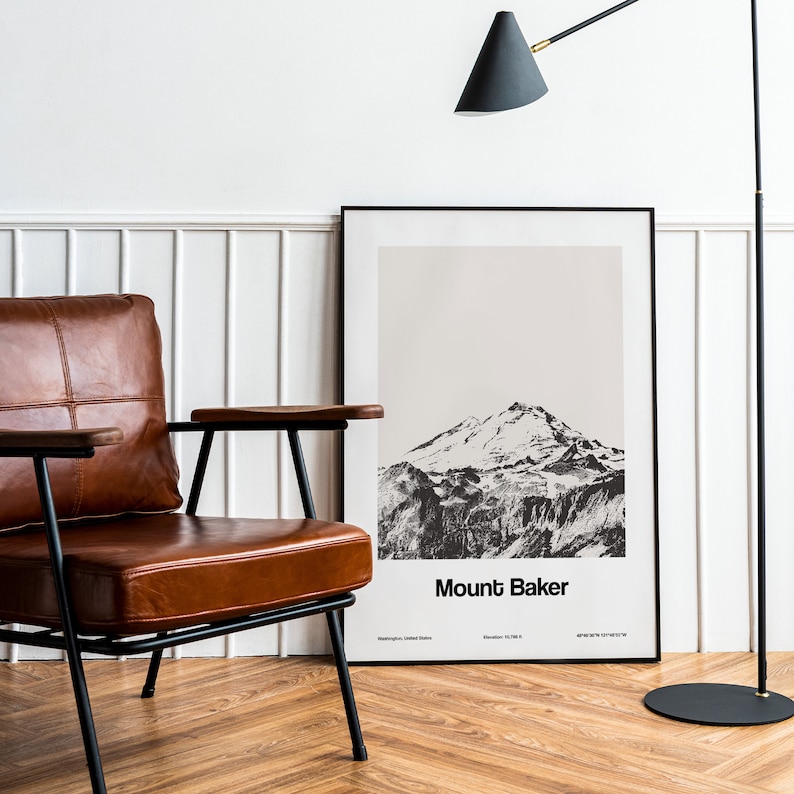Op de afbeelding: Een zwart-witfoto van Mount Baker, een berg in de staat Washington, is ingelijst en hangt aan een witte muur. Een bruine leren fauteuil met zwarte metalen poten staat voor de ingelijste print. Een zwarte staande lamp met een zwarte kap staat op de achtergrond.