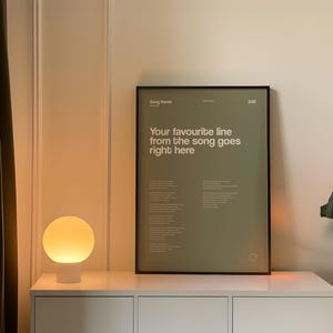 May include: A framed poster with the text "Your favourite line from the song goes right here" in white on a green background. A white lamp with a round, glowing shade sits on a white cabinet. A green plant is visible.