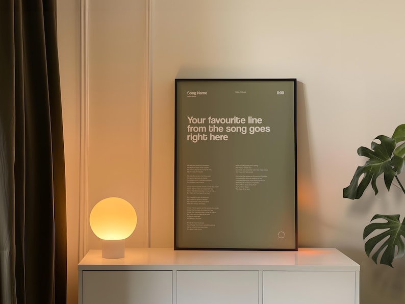 May include: A framed poster with the text "Your favourite line from the song goes right here" in white on a green background. A white lamp with a round, glowing shade sits on a white cabinet. A green plant is visible.