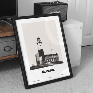 May include: Black and white photograph of Montauk Point Lighthouse in Montauk, New York. The lighthouse is a tall, white structure with a black top. The photo is framed in a black frame.