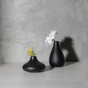 May include: Two black ceramic vases, one tall and narrow, the other short and wide, with a white flower in the tall vase and yellow flowers in the short vase.