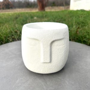 Könnte beinhalten: Weißer Keramiktopf mit einem Gesichtsdesign. Der Topf hat eine strukturierte Oberfläche und eine abgerundete Form.