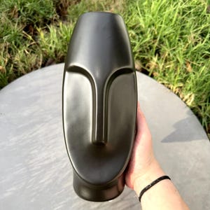 Könnte beinhalten: Eine schwarze Keramikvase in Form eines Gesichts mit glatter, glänzender Oberfläche.
