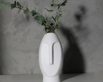 May include: White ceramic vase shaped like a face with a minimalist design. The vase is empty and has a smooth, matte finish.