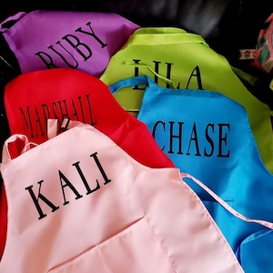 May include: A selection of brightly coloured aprons in purple, green, red, light pink, and blue. Each apron features a pocket and a name printed in black: Ruby, Lila, Marshall, Kali, and Chase. The aprons are made of a smooth fabric.