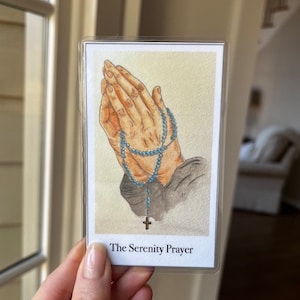 May include: A white card with a watercolor illustration of hands holding a rosary. The card has the text "The Serenity Prayer" printed on the bottom.