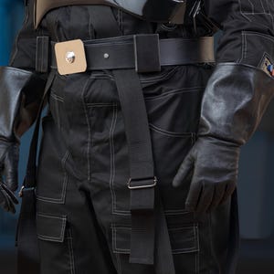 Puede incluir: Traje negro con costuras blancas, con un cinturón ancho con hebilla cuadrada y una correa negra. El atuendo incluye guantes negros y una pieza oscura en el pecho. El disfraz tiene un tema futurista o de ciencia ficción.