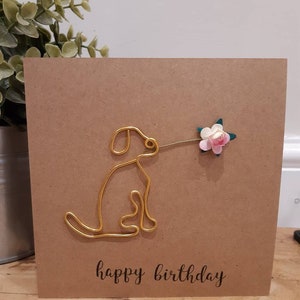 Puede incluir: Una tarjeta marr&oacute;n con un perro de alambre dorado sosteniendo una flor rosa y verde. La tarjeta dice "feliz cumplea&ntilde;os" en letra negra.