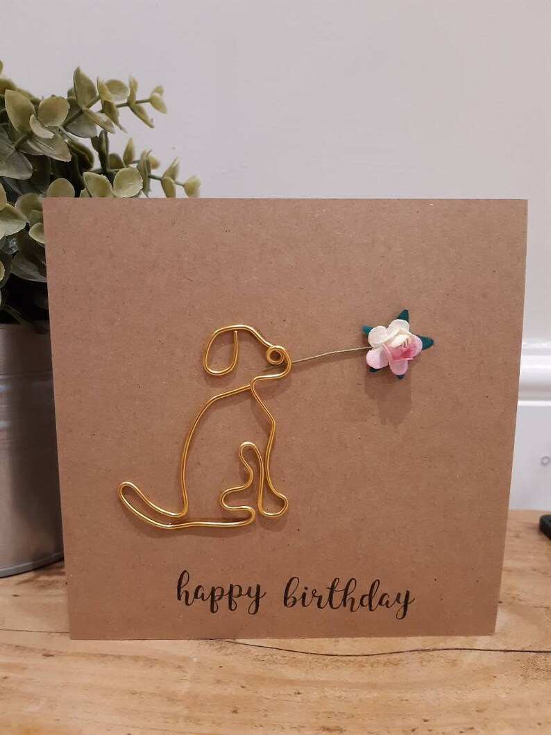 Puede incluir: Una tarjeta marr&oacute;n con un perro de alambre dorado sosteniendo una flor rosa y verde. La tarjeta dice "feliz cumplea&ntilde;os" en letra negra.