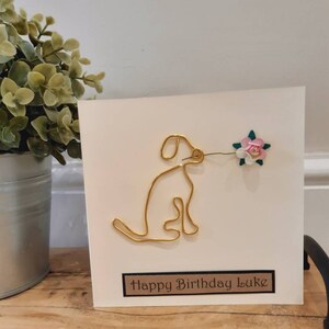 Puede incluir: Una tarjeta blanca con un perro de alambre dorado sosteniendo una flor rosa. La tarjeta dice "Happy Birthday Luke" en una etiqueta marr&oacute;n.