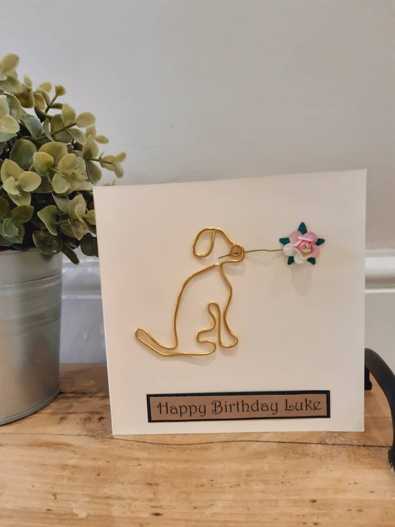 Puede incluir: Una tarjeta blanca con un perro de alambre dorado sosteniendo una flor rosa. La tarjeta dice "Happy Birthday Luke" en una etiqueta marr&oacute;n.