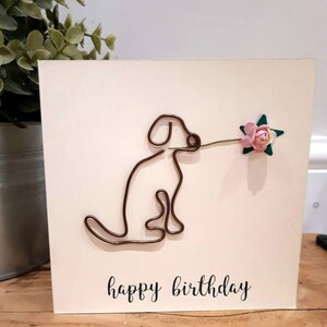 Puede incluir: Una tarjeta blanca con un perro de alambre marr&oacute;n que sostiene una flor rosa con hojas verdes. La tarjeta dice "feliz cumplea&ntilde;os" en cursiva negra.