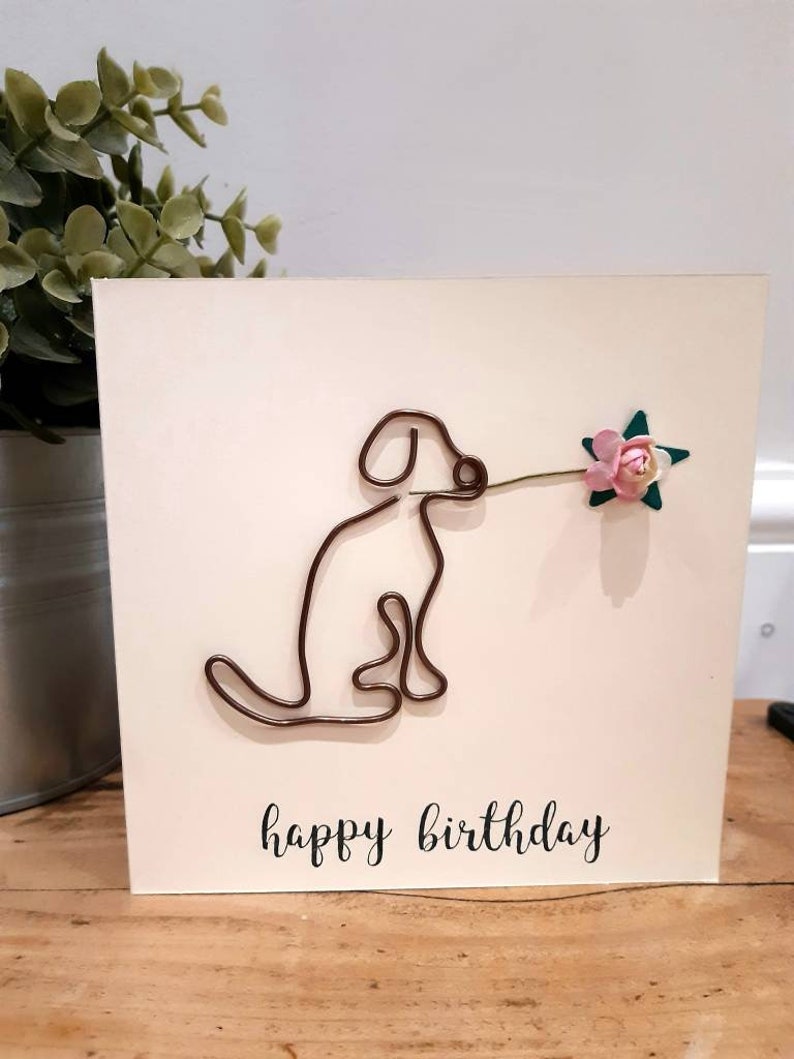 Puede incluir: Una tarjeta blanca con un perro de alambre marr&oacute;n que sostiene una flor rosa con hojas verdes. La tarjeta dice "feliz cumplea&ntilde;os" en cursiva negra.