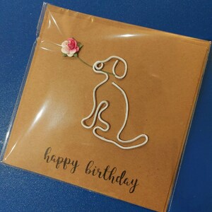 Puede incluir: Una tarjeta marr&oacute;n con un contorno de perro de alambre blanco y una flor de papel rosa y blanco. La tarjeta dice "feliz cumplea&ntilde;os" en letra negra.