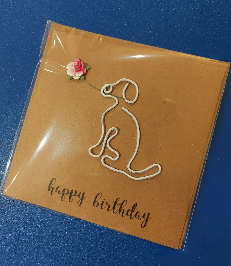 Puede incluir: Una tarjeta marr&oacute;n con un contorno de perro de alambre blanco y una flor de papel rosa y blanco. La tarjeta dice "feliz cumplea&ntilde;os" en letra negra.