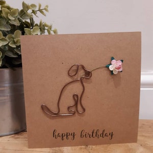 Puede incluir: Una tarjeta marr&oacute;n con una escultura de alambre de un perro sosteniendo una flor rosa. La tarjeta dice "feliz cumplea&ntilde;os" en letras negras.