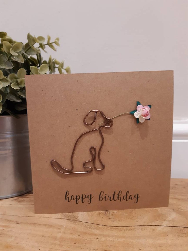 Puede incluir: Una tarjeta marr&oacute;n con una escultura de alambre de un perro sosteniendo una flor rosa. La tarjeta dice "feliz cumplea&ntilde;os" en letras negras.