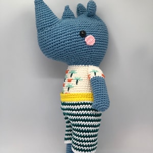 May include: A blue crocheted rhinoceros wearing a white, yellow, and green striped outfit. The rhinoceros has a black eye and a pink nose.