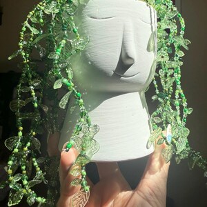 May include: A white ceramic head-shaped planter featuring a minimalist facial design. The planter is adorned with cascading strands of green glass beads and translucent green leaf-shaped charms that drape over the sides, mimicking trailing vines.